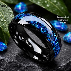Könnte beinhalten: Ein schwarzer Ring mit einer zentralen Einlage aus zerkleinerten, irisierenden blauen und grünen Steinfragmenten. Die Innenseite des Rings ist mit dem Text My Person graviert, und das Bild enthält den Text Personalized Engraving Inside.
