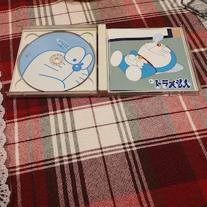 May include: Two Doraemon CD cases are open on a red and white plaid surface. One case shows a blue CD with a Doraemon illustration. The other case shows Doraemon lying down. Japanese text is visible.