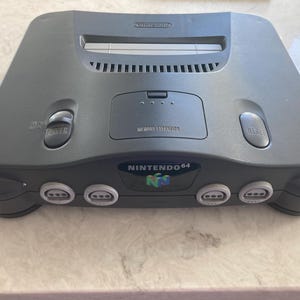 May include: A grey Nintendo 64 video game console. The console features a power button, reset button, and four controller ports. The Nintendo 64 logo is displayed on the front.
