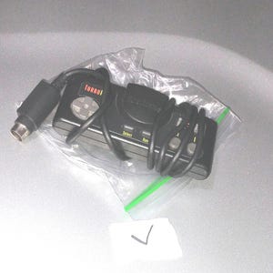 Puede incluir: Un mando TurboPad negro con una cruceta gris y texto rojo. El mando está envuelto en una bolsa de plástico transparente con cremallera verde. Tiene un cable negro y un conector plateado.