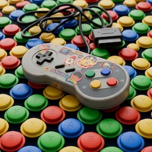 May include: A gray video game controller with a black directional pad and four colored action buttons, decorated with various small stickers, resting on a surface covered in a repeating pattern of red, blue, green, and yellow circular buttons.