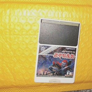 Peut inclure: Une cartouche de jeu vidéo rectangulaire avec un dessus noir et un graphisme coloré d'un guerrier ninja. La cartouche est étiquetée "namcot" et "HuCARD" et est posée sur un fond jaune à bulles.