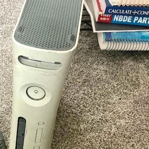 May include: A white Xbox 360 console with a gray top panel and a power button. The console is slightly angled, showing its front panel with a disc drive slot and various buttons. A stack of books is visible in the background.