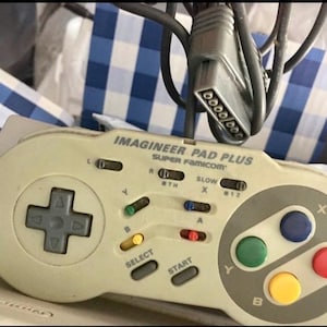 Puede incluir: Un mando Imagineer Pad Plus Super Famicom vintage en blanco roto con una cruceta gris, botones de colores y un cable gris. El mando tiene el texto "IMAGINEER PAD PLUS SUPER Famicom".