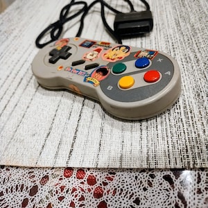 May include: A gray Super Nintendo controller with colorful buttons and a black cord. The controller has stickers on it with cartoon characters and the text "Go for it!!"