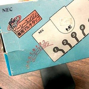 Puede incluir: Un dispositivo multi-tap PC Engine vintage en su caja original. La caja es de color turquesa con rayas blancas y presenta texto japonés y el logotipo de NEC. El multi-tap tiene cinco puertos de controlador numerados del 1 al 5.