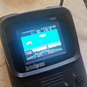 May include: Black Turbo Express handheld game console with a screen displaying a racing game. The game features a road, car, and the Eiffel Tower. The console has the text "Turbo Express" and "TurboGrafx 16" on it.