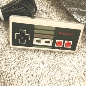 May include: A classic Nintendo controller with a black directional pad, gray select and start buttons, and red A and B buttons. The controller has a white and black color scheme with the Nintendo logo.