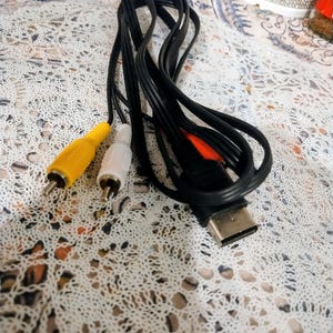 May include: Black AV cable with yellow, white, and red RCA connectors. The cable has a rectangular connector on one end. The cable is on a white lace surface.