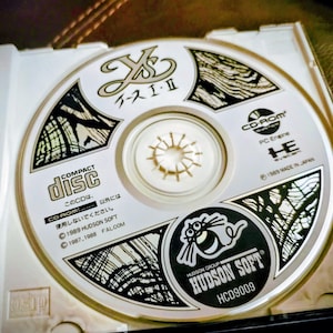 May include: A compact disc for the video game Ys I & II, in a white case. The disc features Japanese text and a graphic of a cartoon character. The disc is labeled "HUDSON SOFT" and "HCD9009".