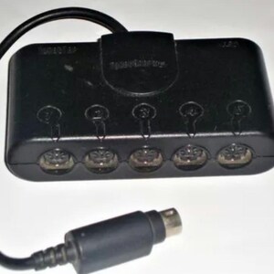 Puede incluir: Adaptador negro para mandos de GameCube con cinco puertos y un cable negro. El adaptador tiene el texto "GameTap" y "speedrunner" impreso. El adaptador está diseñado para conectar varios mandos a una consola GameCube.