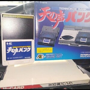 Peut inclure: Une cartouche de jeu HuCARD blanche avec une étiquette bleue portant du texte japonais et le titre "TENNOKOE BANK". Une boîte de jeu assortie est derrière, avec le même titre en rouge et du texte japonais. La boîte montre des consoles et des cartouches de jeu.