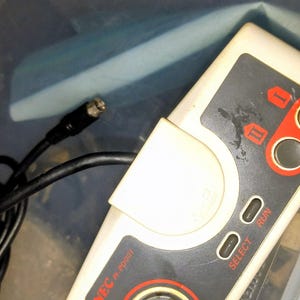 Puede incluir: Un mando de videojuegos vintage NEC PI-PD001. El mando es de color blanco roto y gris con detalles en rojo. Cuenta con una cruceta direccional, botones de selección y ejecución, y dos botones de acción. Un cable negro con un conector sale del mando.