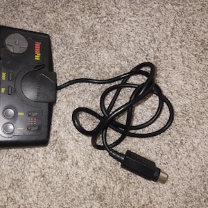 Puede incluir: Mando Turbo Pro negro con cruceta, botones y un cable negro largo. El mando tiene el texto "Turbo Pro" en rojo. El mando está diseñado para jugar.