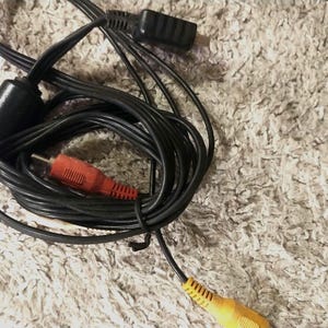 May include: A black AV cable with a red RCA connector and a yellow RCA connector. The cable is coiled and has a black power adapter. The connectors are visible against a textured, light-colored background.
