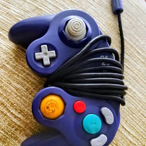 May include: A purple video game controller with a black cord. The controller has a directional pad, a joystick, and several colored buttons, including red, green, and yellow. The controller is designed for gaming.
