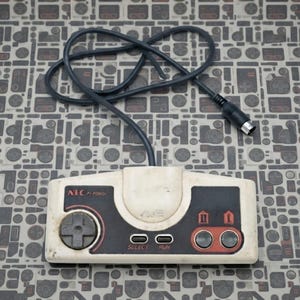 May include: A white and black NEC PI-PD001 video game controller featuring a directional pad, two action buttons labeled I and II, and select and run buttons. The controller includes a black wired cable with a circular connector plug at the end.
