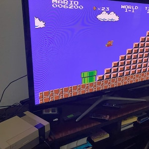 May include: A television screen shows a classic video game scene with pixelated graphics. The game features a blue sky, brown blocks, and a green pipe. A retro game console sits below the screen on a dark wooden surface.
