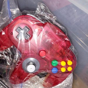 May include: A transparent red Nintendo 64 controller with a gray directional pad, red, green, blue, and yellow buttons. The controller is in a clear plastic bag.