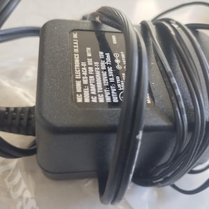 May include: Black AC adapter for NEC TurboGrafx-16, with a black power cord wrapped around it. The adapter has the text "NEC HOME ELECTRONICS (U.S.A.) INC" and other technical specifications printed on it.