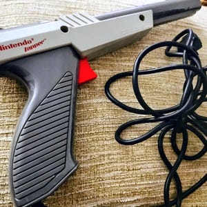 May include: A gray and black Nintendo Zapper light gun with a red trigger and a black cord. The gun has the text "©1985 Nintendo Zapper" printed on the side. The gun is a retro gaming accessory.