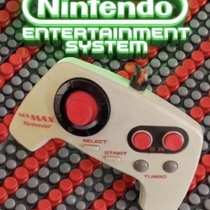 Könnte beinhalten: Ein beigefarbener Nintendo NES Max Videospiel-Controller mit einem kreisförmigen Steuerkreuz mit roter Mitte, zwei roten A- und B-Tasten, zwei grauen Turbo-Tasten und roten Beschriftungen. Der Controller trägt den Schriftzug NES MAX Nintendo.