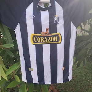 Può includere: Una maglia da calcio a righe verticali bianche e nere con maniche nere. La parte anteriore mostra la parola "CORAZON" in un emblema giallo e marrone, insieme a loghi e stemmi della squadra.