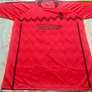 May include: A red short-sleeved jersey with black trim and accents. The word "MEXICO" is printed across the chest in dark blue. A black zig-zag pattern is above the word "MEXICO".