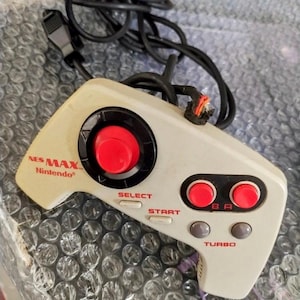 May include: Off-white NES Max controller with a large red circular button and several smaller red and gray buttons. The controller has the text "NES MAX Nintendo" printed on it. A black cable is attached.