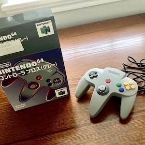 May include: A grey Nintendo 64 video game controller with a black wired cord and its original retail box. The box features the Nintendo 64 logo, the text NINTENDO 64, and a graphic illustration of the controller with Japanese text characters.
