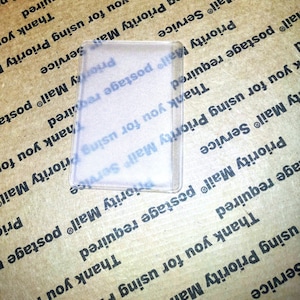 Puede incluir: Un tarjetero rectangular de plástico transparente. El soporte es transparente, lo que permite ver la tarjeta en su interior. El fondo es una superficie de cartón marrón con texto impreso, incluyendo "Priority Mail Service" y "Thank you for using Priority Mail".