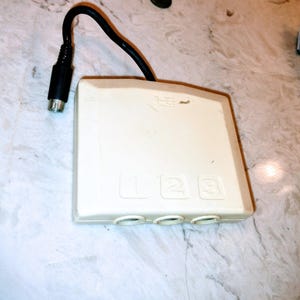 Puede incluir: Dispositivo electrónico blanquecino con tres botones numerados (1, 2, 3) y tres puertos circulares. Un cable negro con un conector se extiende desde la esquina superior izquierda. El dispositivo está sobre una superficie con vetas de mármol.