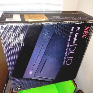 May include: A black box for a PC Engine Duo video game console. The box features a graphic of the console in dark blue and purple, with the text "PC Engine Duo" and "PC Engine with SUPER CD-ROM²". The box is stacked on a green box.
