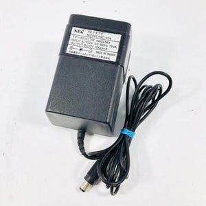 May include: A black NEC AC adapter, model PAD-124, with a black cable secured by a blue rubber band. The adapter has a white label with Japanese text and technical specifications.
