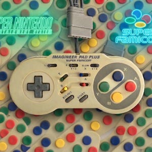 Puede incluir: Un mando de juego Imagineer Pad Plus de color blanco roto para Super Famicom, con una cruceta gris, cuatro botones de acción de colores etiquetados como A, B, X e Y, y un cable de conexión gris. El texto Super Nintendo Entertainment System y Super Famicom aparece en azul neón.