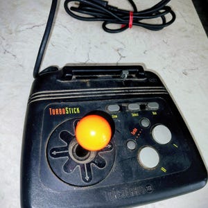 Puede incluir: Mando TurboStick negro con un joystick naranja y un cable negro. El mando tiene botones con las etiquetas "Slow", "Select" y "Run". El texto "TurboGrafx 16" está impreso en el mando.