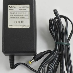 May include: Black NEC AC adapter, model PAD-129, with a label displaying technical specifications. The adapter has a black power cord bundled with a green band. The label reads "INPUT: AC100V 50-60Hz 20VA OUTPUT: DC9V 1000mA".