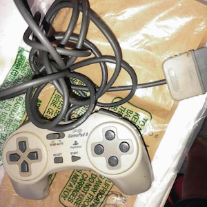May include: A vintage PlayStation GamePad 8 controller in a light gray color. The controller has a directional pad, four action buttons, and start/select buttons. The controller is connected to a gray cable.