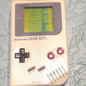 May include: A vintage Nintendo Game Boy handheld console in a cream color. The screen displays a green game menu. Features include a directional pad, pink buttons, and the text "Nintendo GAME BOY".