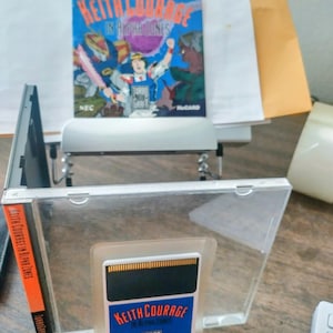May include: A clear plastic case containing a video game cartridge and a game box. The cartridge has the text "KEITH COURAGE" and "TURBO GRAFX" on a blue background. The box art features the title "KEITH COURAGE IN ALPHA ZONES".