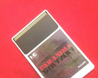 May include: A white game cartridge with a black and silver design. The text "PARANOIA" is printed in red on the cartridge. The text "HuCARD" is printed in black on the cartridge.