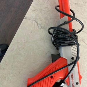 May include: A red and gray Nintendo Zapper light gun with a black cable. The gun has a red handle and barrel, with a gray body and the Nintendo logo. The cable is coiled around the gun.