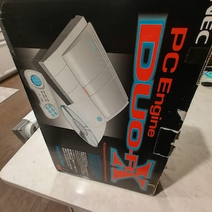 May include: A black and blue PC Engine Duo-R game console box. The box features a graphic of the console and controller, with the words "PC Engine Duo-R" in red and blue. The box is slightly damaged.
