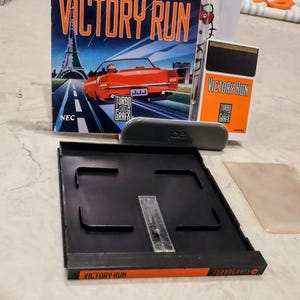 Pode incluir: Um conjunto de itens de videogame retrô, incluindo o jogo "Victory Run". A caixa do jogo mostra um carro vermelho em uma estrada com a Torre Eiffel ao fundo. Inclui um cartucho de jogo, uma caixa e outros acessórios.