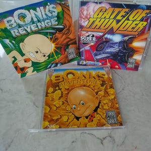 Op de afbeelding: Drie TurboGrafx-16 videogame-cases. De games zijn "Bonk's Revenge", "Gate of Thunder" en "Bonk's Adventure". De "Bonk's Revenge"-case heeft een stripfiguur op een jungle-achtergrond. "Gate of Thunder" heeft een ruimteschip-afbeelding. "Bonk's Adventure" heeft een stripfiguur op een bruine achtergrond.