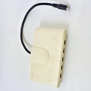 Puede incluir: Un dispositivo electrónico de color crema con un cable y conector negros. El dispositivo tiene cinco puertos numerados y una pequeña tapa con bisagras. El cable está conectado a la parte superior del dispositivo.