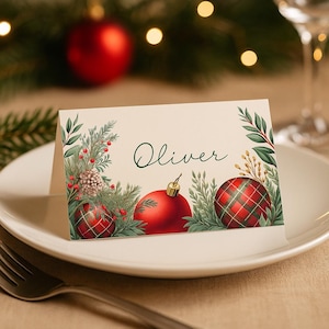 Peut inclure: Une carte de place crème avec le nom "Oliver" en écriture cursive, encadrée de verdure festive et d'ornements rouges. La carte présente une boule de Noël rouge et deux ornements à carreaux. La carte est sur une assiette blanche.