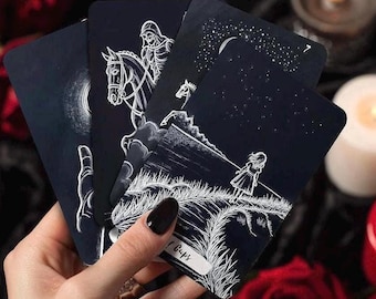 May include: A hand holds a fan of tarot cards with dark blue backgrounds and white illustrations. The cards depict scenes of a skeleton on horseback, a girl in a field, and a crescent moon. The cards are part of a tarot deck.
