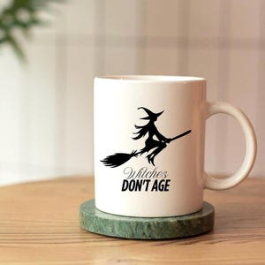 Può includere: Tazza in ceramica bianca con silhouette nera di una strega su una scopa e la scritta "Witches DON'T AGE". La tazza è su un sottobicchiere di marmo verde. Un regalo divertente per Halloween o per gli amanti delle streghe.