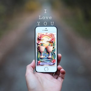 May include: A smartphone displays a colorful illustration of two cups, flowers, and a sun, with the text "Two of Cups." Above the phone, the words "I Love You" are written. The phone is held against a blurred background.